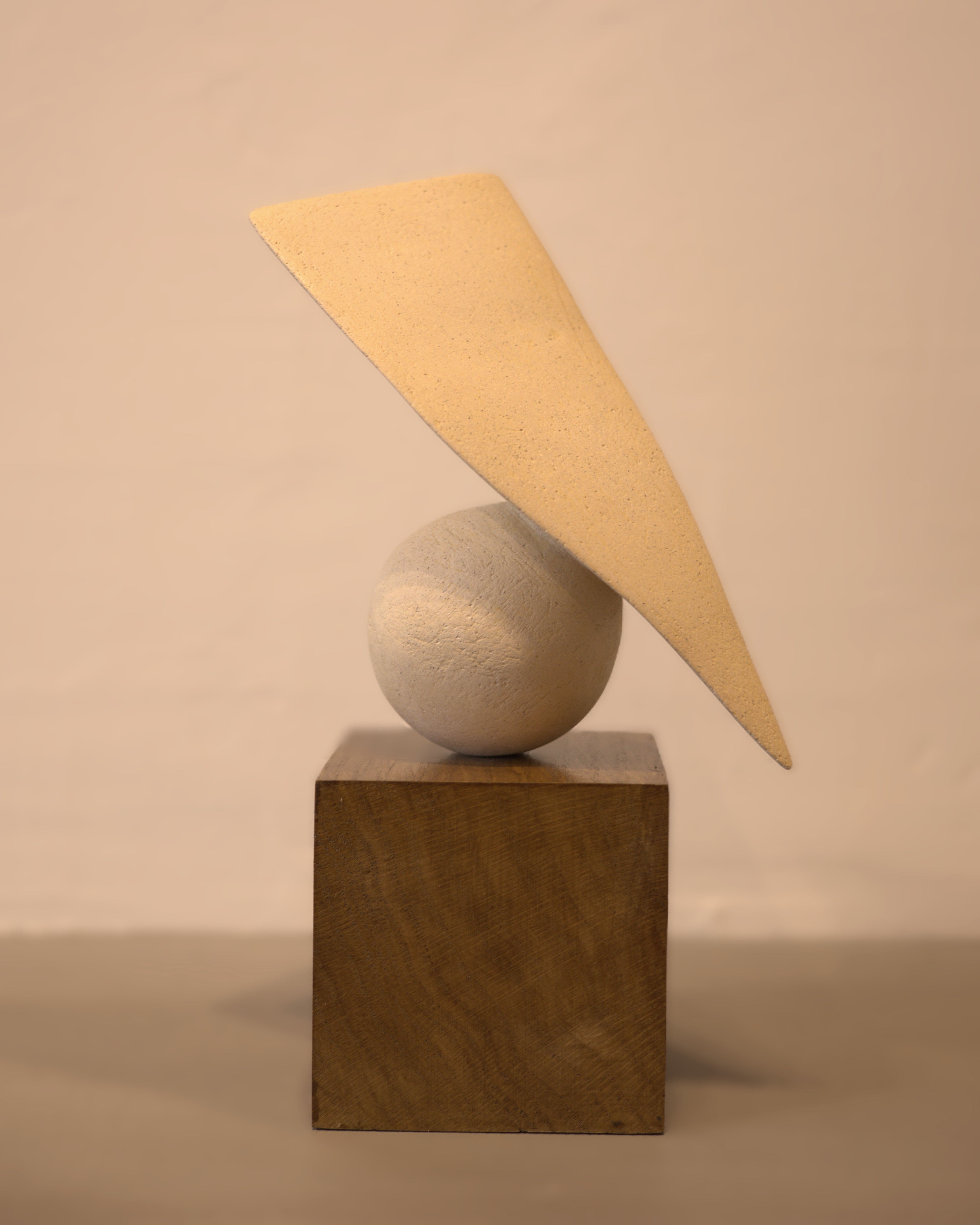 White ceramic sphere and angled feather balanced on a warm wood cube; three-element geometric sculpture on neutral ground.